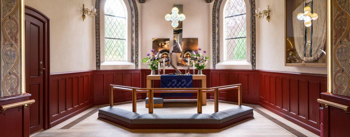 Interiør af en kirke med alter, blomster og farverige vinduer i en lys og indbydende atmosfære.