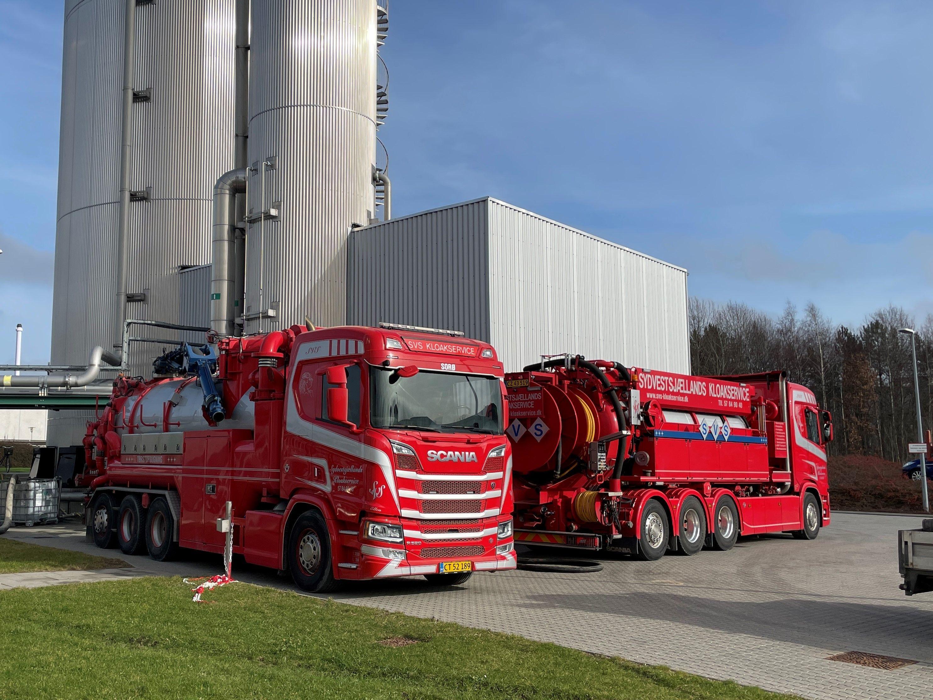 To store og røde lastbiler fra Scania parkeret ved en industriel bygning med grå vægge.