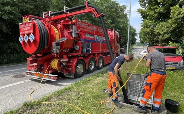 Toiletvogn i rødt ved en vej, mens to arbejdere rengør en brønd med slanger og udstyr.