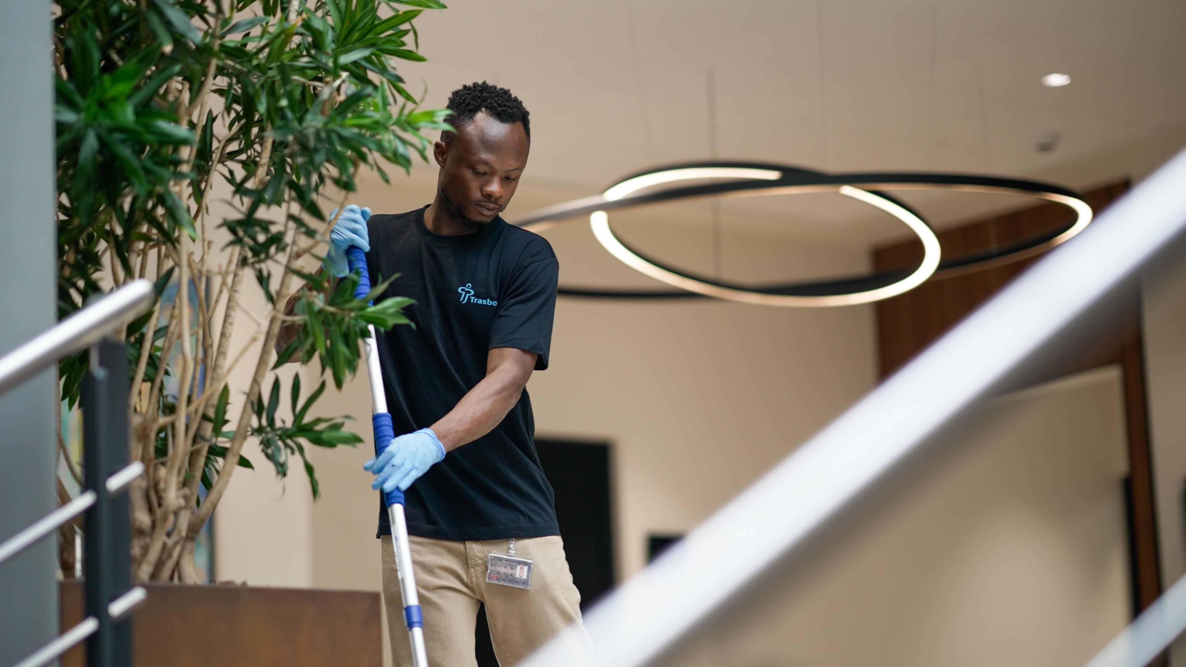 En mand i sort t-shirt og handsker, der mopper gulvet i en moderne bygning med planter og belysning.