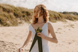 En kvinde i en hvid kjole står på stranden med bølgerne i baggrunden og holder en grøn sløjfe.