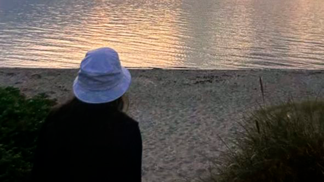 En person med en hvid hat står ved stranden og ser på vandet ved solnedgang.
