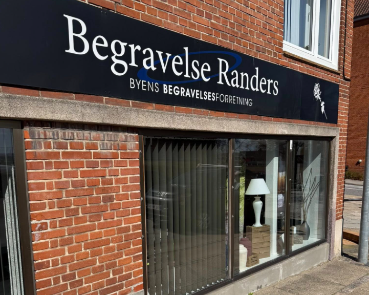 Facade af Begravelse Randers med skilt og vindue, der viser en lampe og indretning.