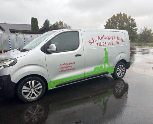S.E. Anlægsgartner bil med logo og kontaktinformation, parkeret på en våd vej.