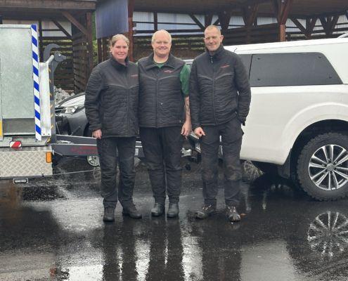 Tre personer står sammen foran en bil på en regnfuld dag, iført sorte jakker og arbejdstøj.