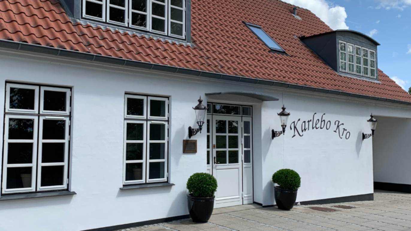 En hvid bygning med rødt tag og store vinduer, der viser indgangen til Kærbo Kro.