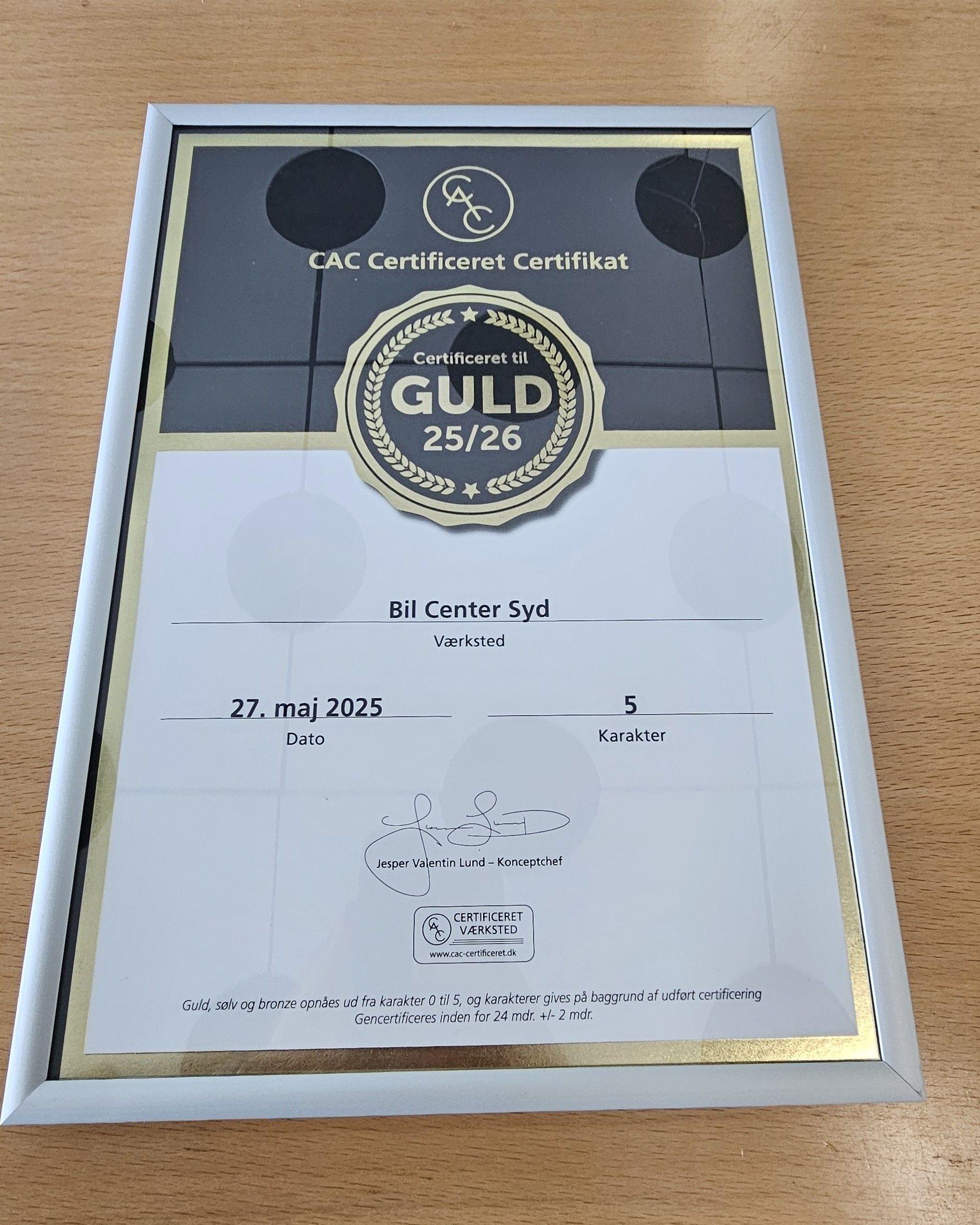 CAC certifikat i ramme med teksten "GULD 25/26" og datoen "27. maj 2025" på en træoverflade.
