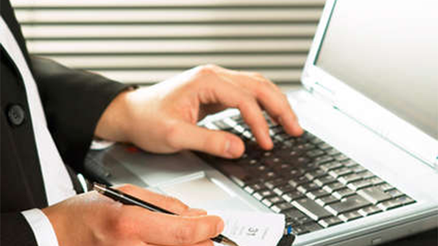 En mand i jakkesæt skriver på en bærbar computer med en pen i hånden.