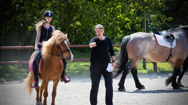 En rytter på en pony modtager instruktioner fra en træner på en rideskole i solrigt vejr.