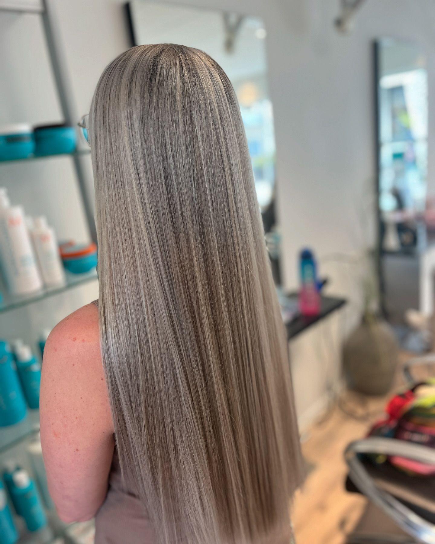 Langt, glat hår i en lys grå farve, set bagfra i en frisørsalon med produkter i baggrunden.