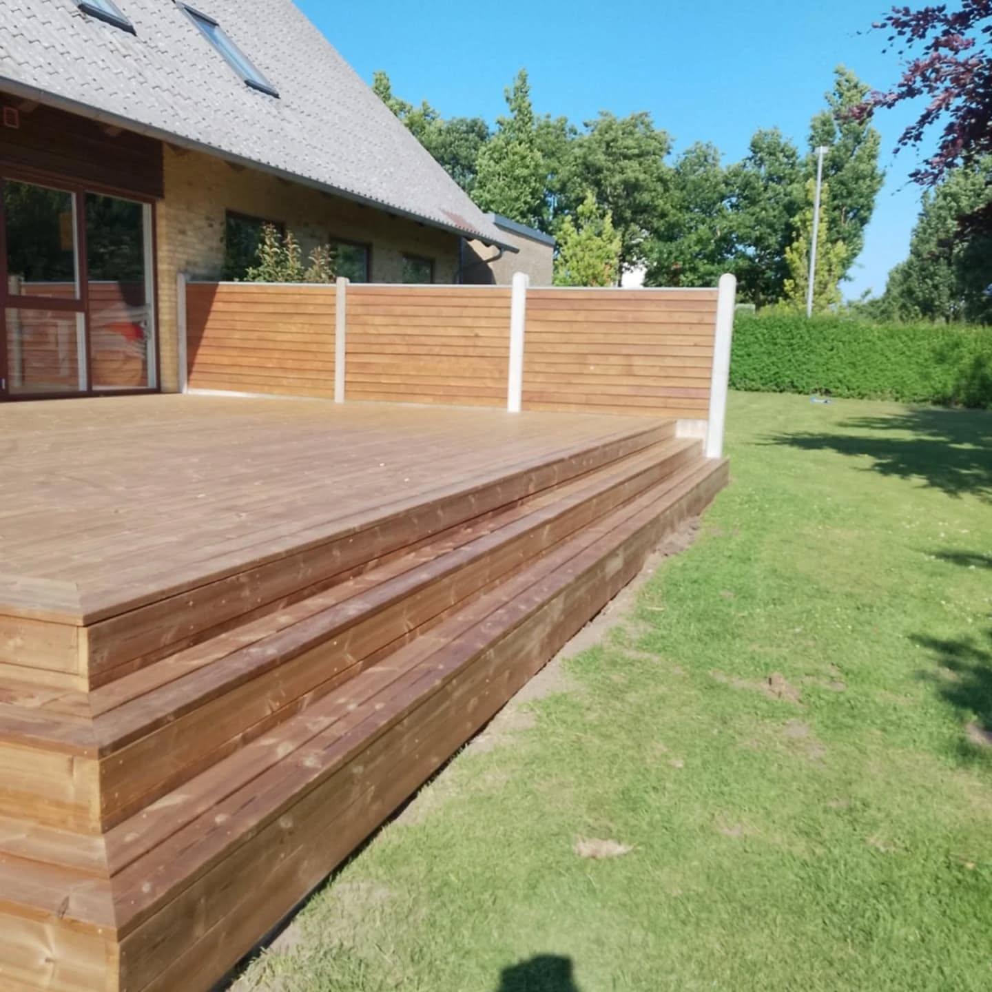 Terrasse i træ med trin og hvidt rækværk ved en husfacade, omgivet af grøn græsplæne og træer.