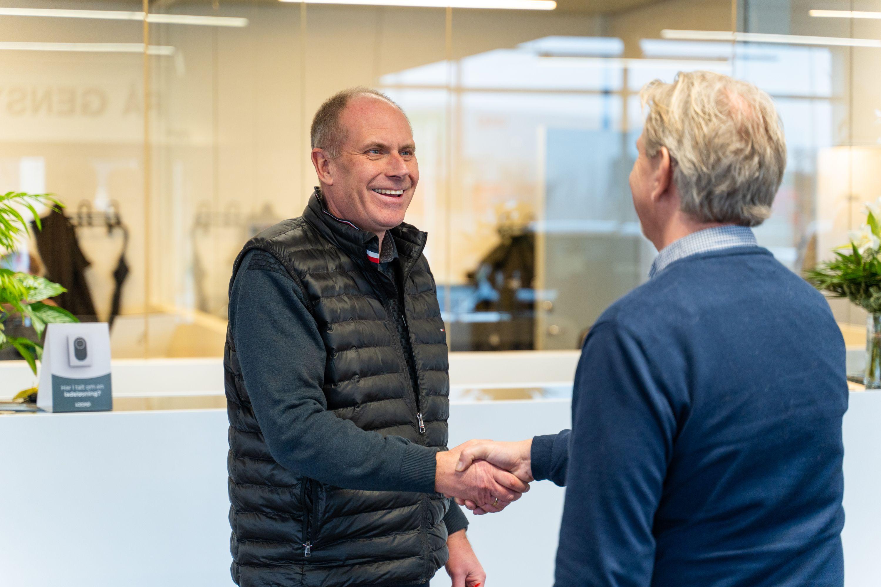 To mænd, der håndterer et håndtryk i et moderne kontormiljø med planter og en reception i baggrunden.
