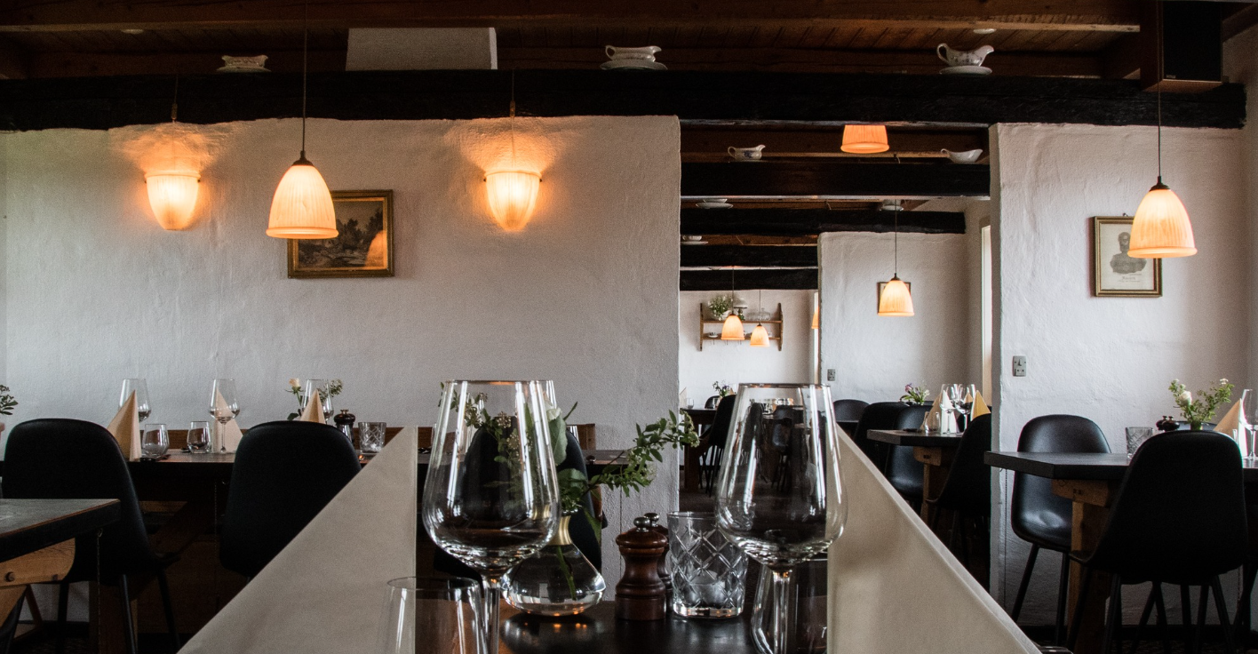 Indendørs restaurant med elegante borde, glas og blomsterdekorationer, oplyst af bløde lamper.