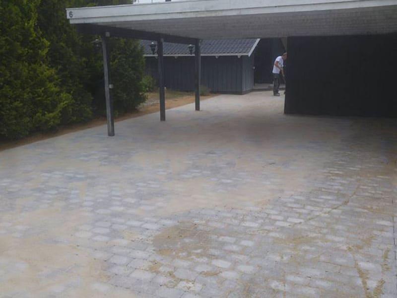 En grusbelagt indkørsel under en carport med træer og bygninger i baggrunden.