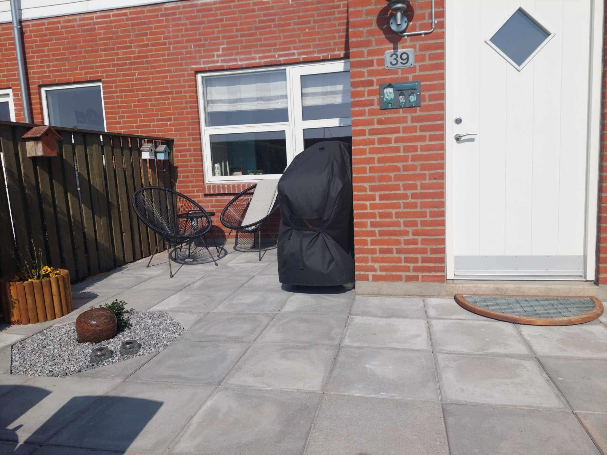 En terrasse med fliser, en sort grill, to stole og en blomsterkasse ved en hvid dør.