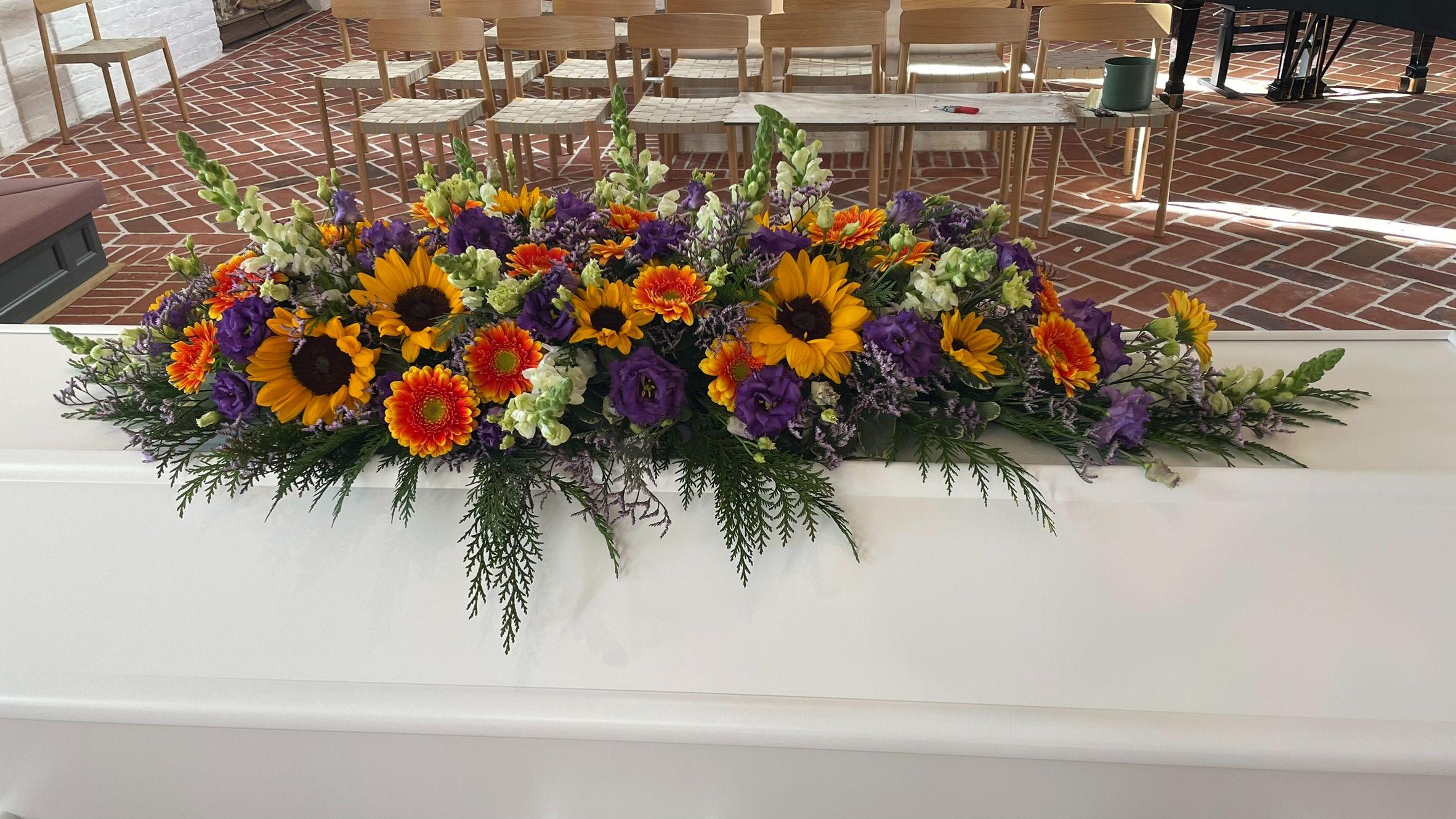 En smuk blomsterkrans med solsikker, gerberaer og lilla blomster på en hvid kiste i en kirke.