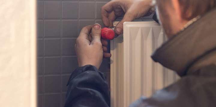 En håndværker justerer en radiator med en rød ventilnøgle i et badeværelse med fliser.