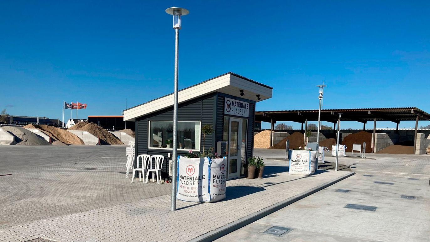 En lille informationsbod ved en parkeringsplads med blå himmel og sandbunker i baggrunden.