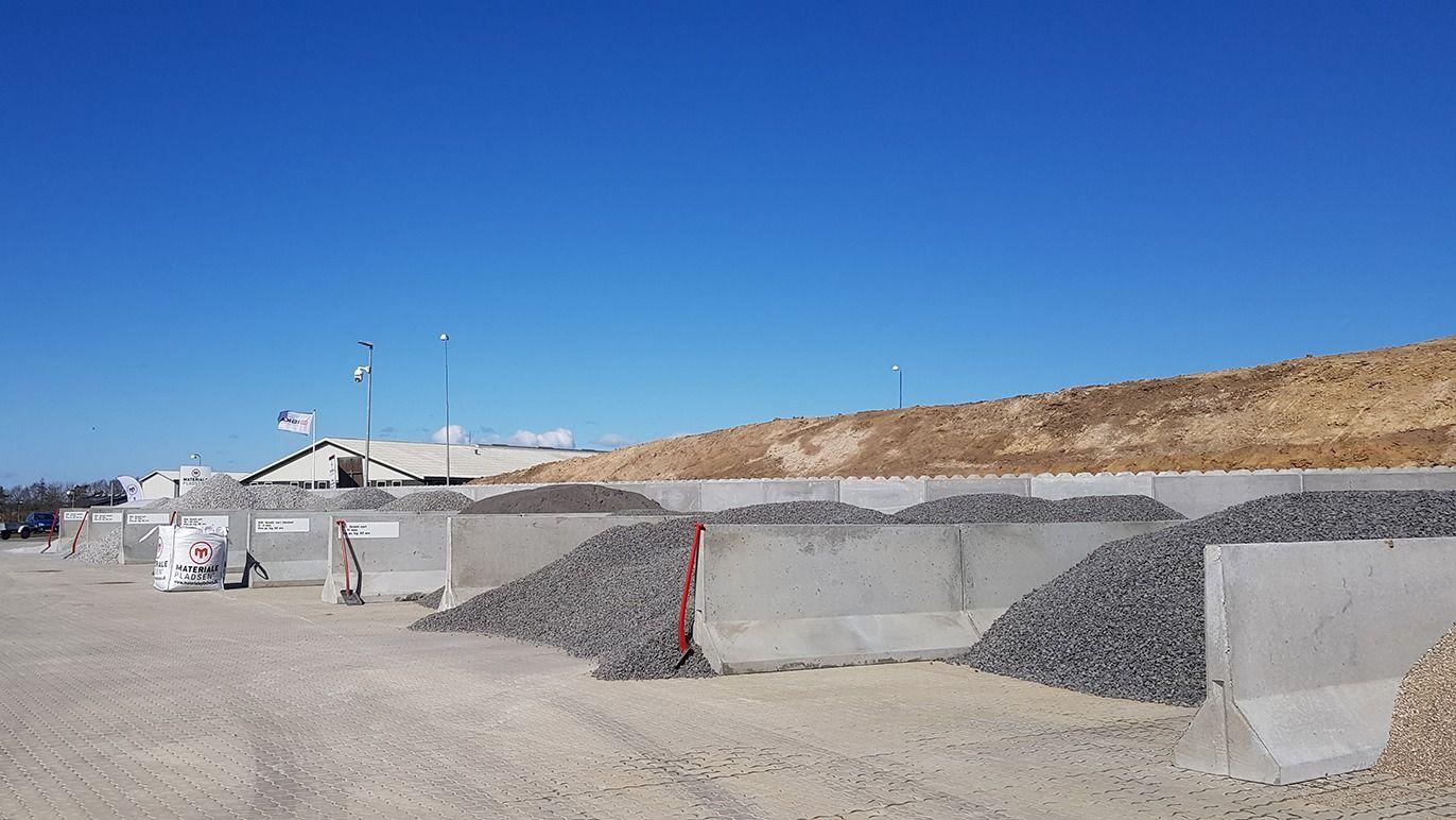 Grus- og stenlager med forskellige materialer i betonbeholdere under en klar blå himmel.