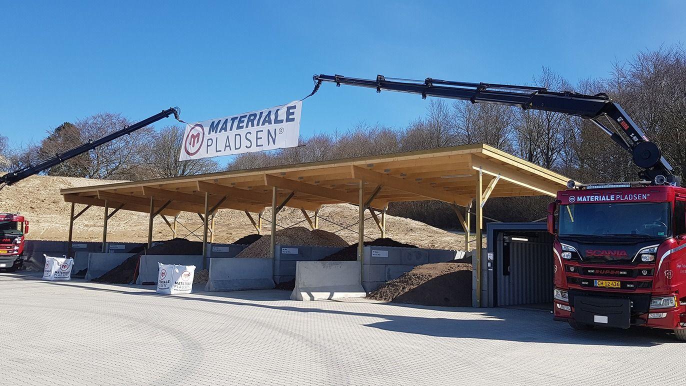Materialeplads med overdækning, lastbiler og kraner, der håndterer byggematerialer under en blå himmel.