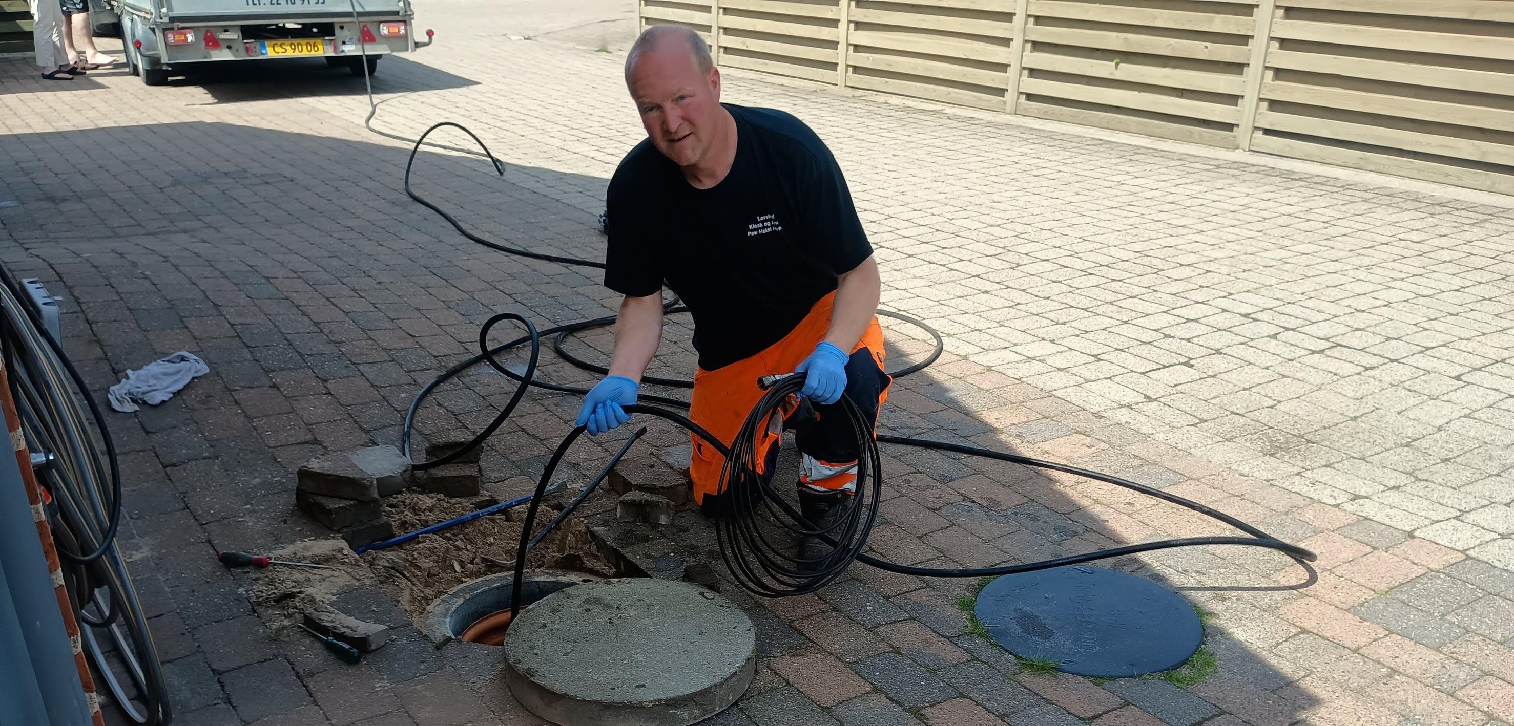 En mand i orange bukser arbejder med en slamsuger ved en brønd på en brostenbelagt vej.