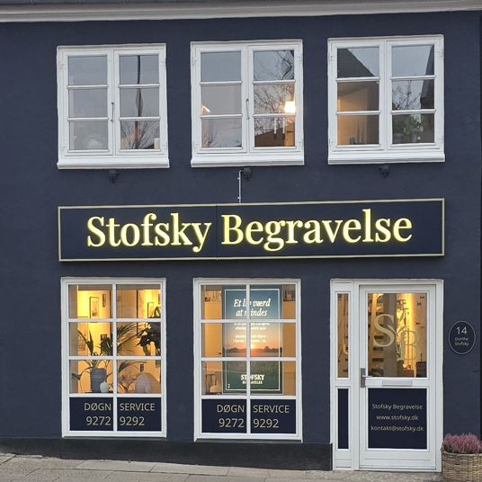 Butik med skilt for Stofsky Begravelse, vinduer med lys og serviceinformation i en mørkeblå bygning.