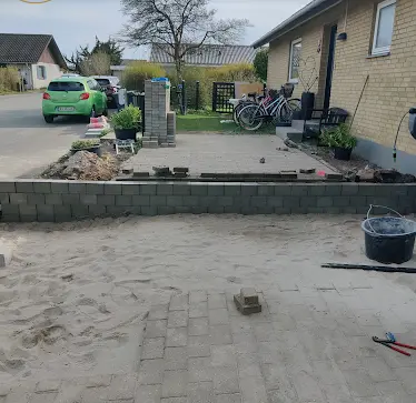 Billede af en gård med en nybygget mur, sandbund og en grøn bil parkeret i baggrunden. Cykler står ved siden af huset.