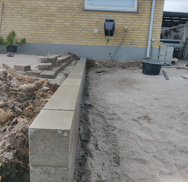Murstenstablet langs en sandet grund ved en bygning, med værktøj og materialer i baggrunden.