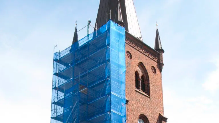 Kirketårn med stillads dækket af blå plastik, der viser renovering af bygningens facade.