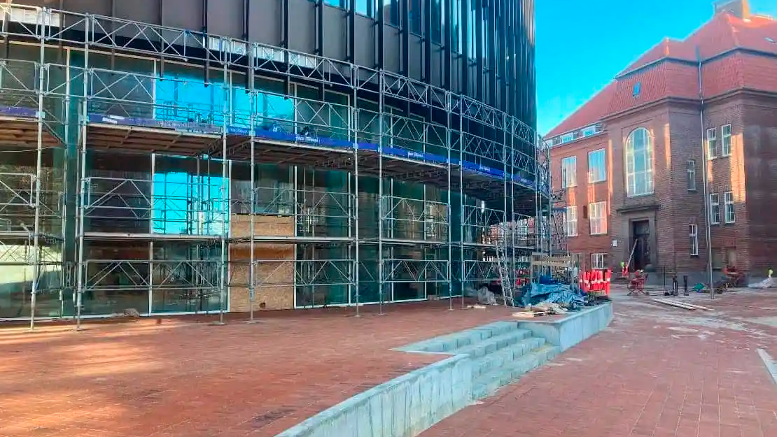 Byggeri med stillads og glasfacade, der viser en moderne bygning ved siden af en ældre murstensbygning.