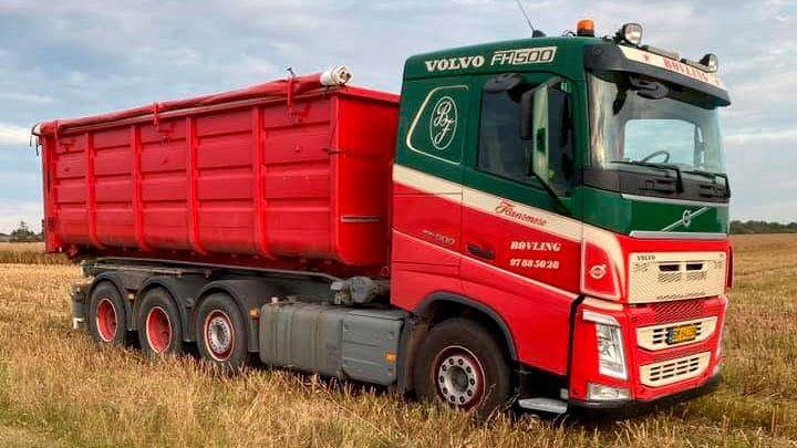 En rød Volvo lastbil med en stor container står på en mark under en blå himmel.