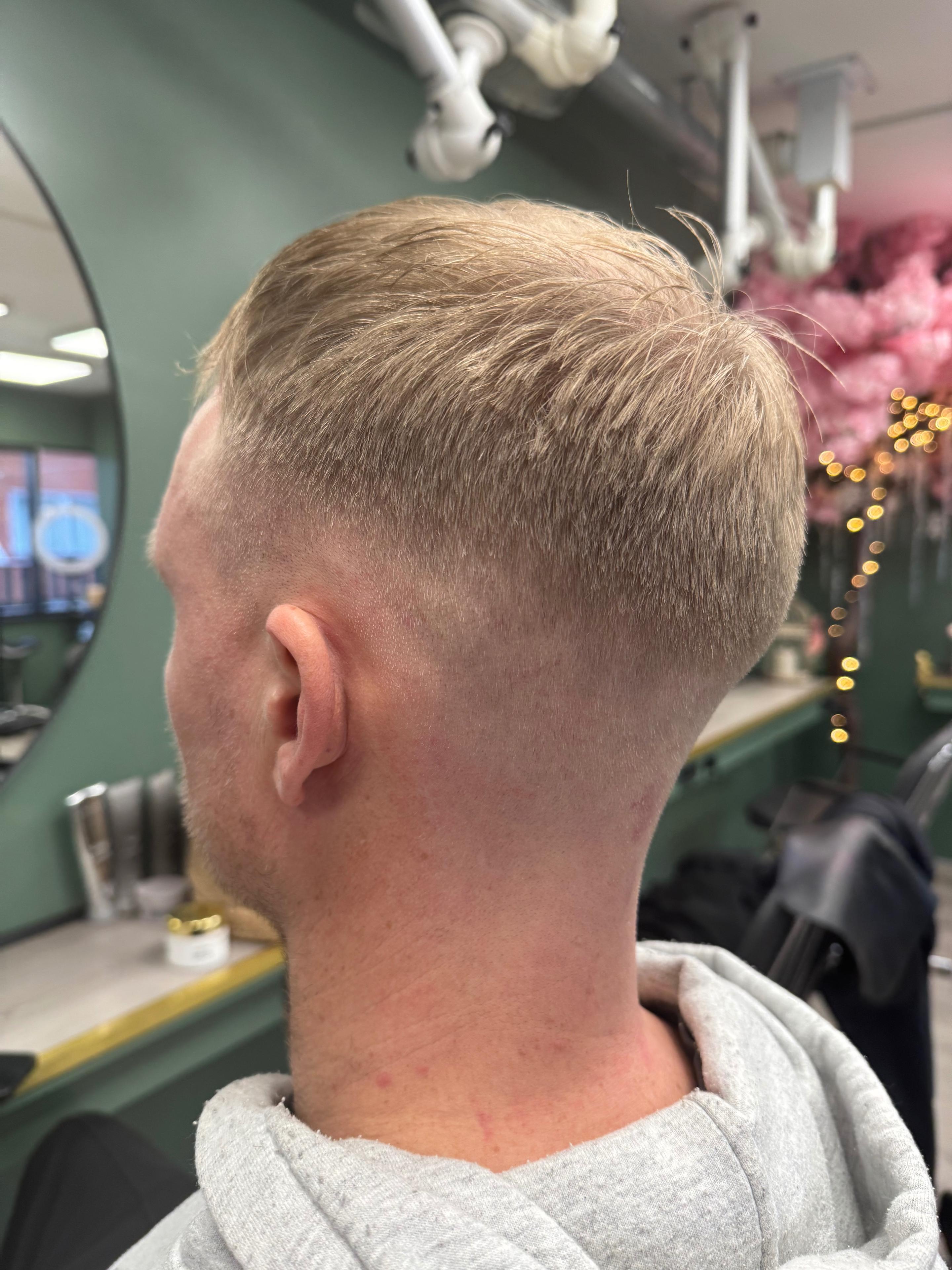 En mand med kort hår og fade haircut står i en frisørsalon med grønne vægge og spejl.