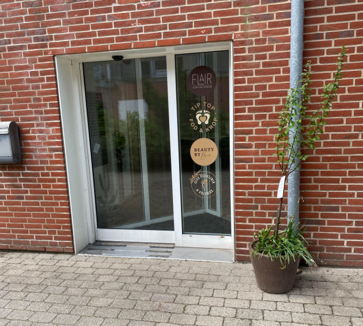 Indgangsdør til en bygning med glasdør og murstensvæg, prydet med skilte og en potteplante ved siden af.