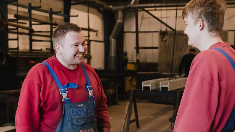 To mænd i arbejdstøj smiler og snakker sammen i en værksted med metaludstyr i baggrunden.