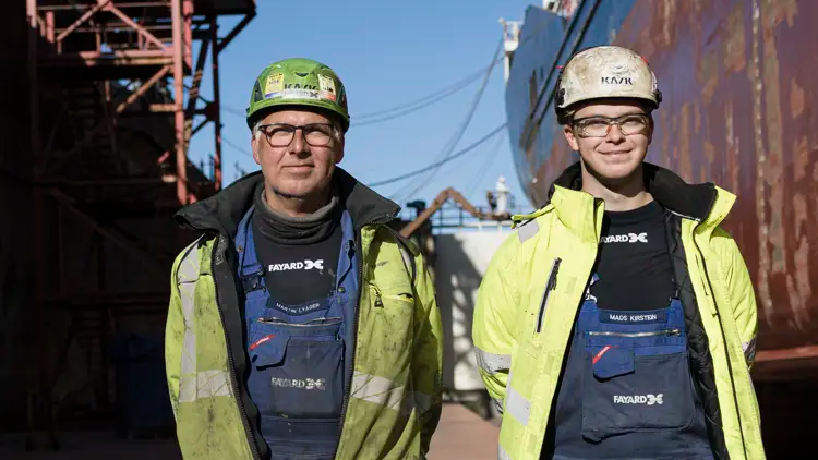 To arbejdere iført sikkerhedshjelme og arbejdstøj står foran et skib i en havn.