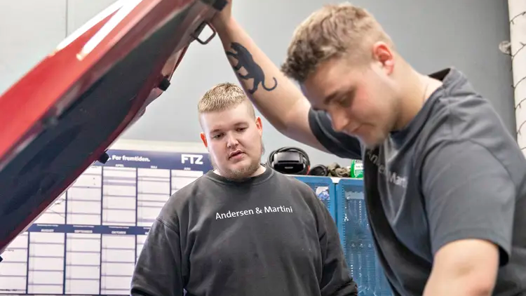 To unge mænd arbejder sammen på en bilreparation i et værksted, hvor den ene ser på motoren under motorhjelmen.