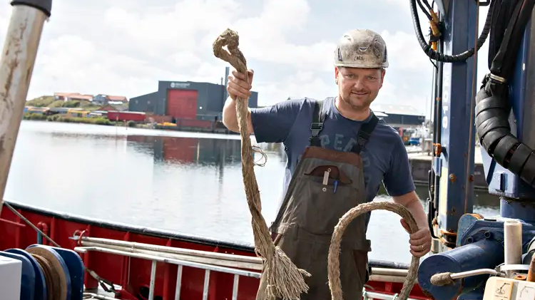 En mand i hjelm holder to reb på en båd ved havnen med bygninger i baggrunden.