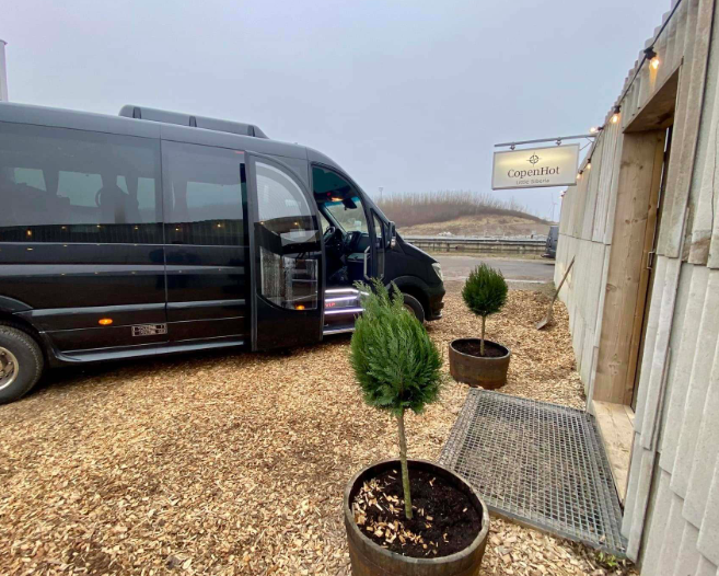En sort minibus parkeret ved en restaurant med små træer i potter og grusbelægning.