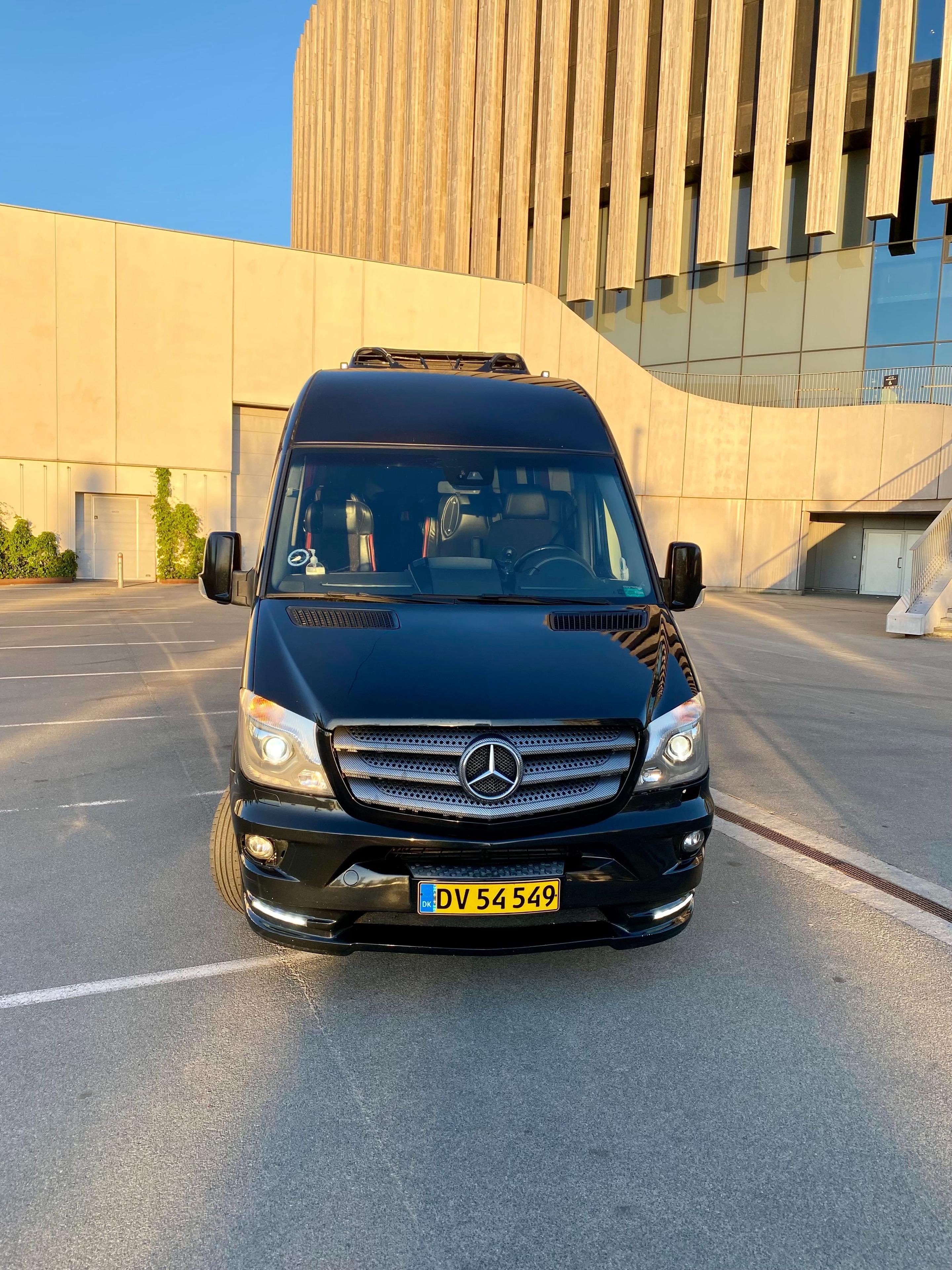 En sort Mercedes varevogn parkeret foran en moderne bygning med glasfacade.