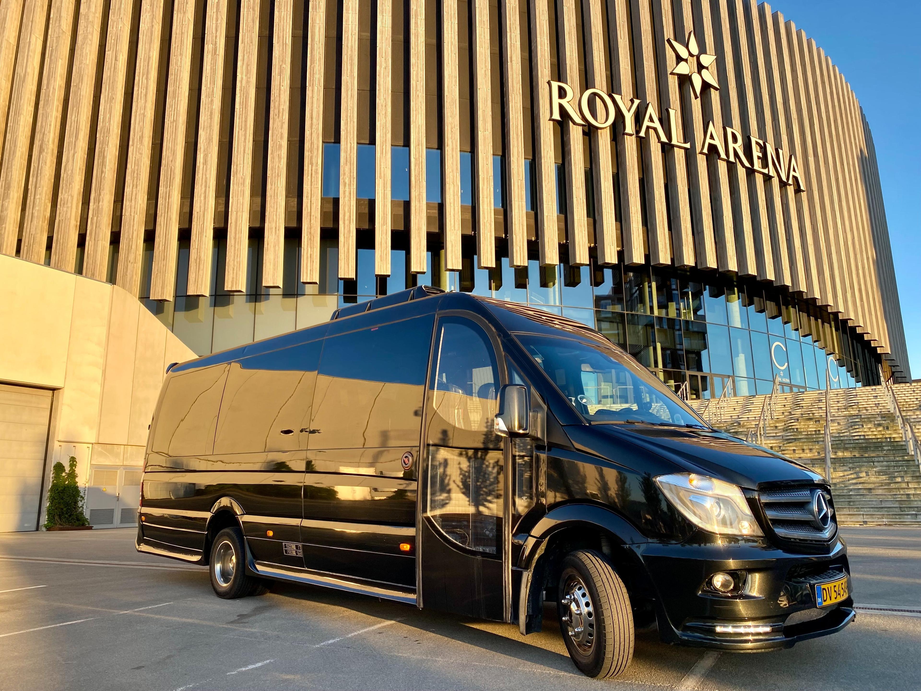 En sort Mercedes-bus parkeret foran Royal Arena med moderne arkitektur i baggrunden.