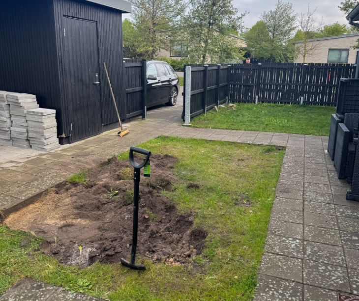 En have med en gravet hule, en skovl, græsplæne og en sort garage i baggrunden.