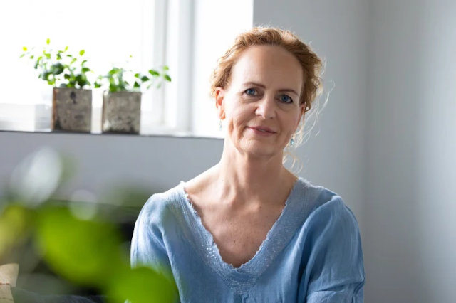 Kvinde med lys skjorte sidder i stue med planter i vinduet og smiler til kameraet.
