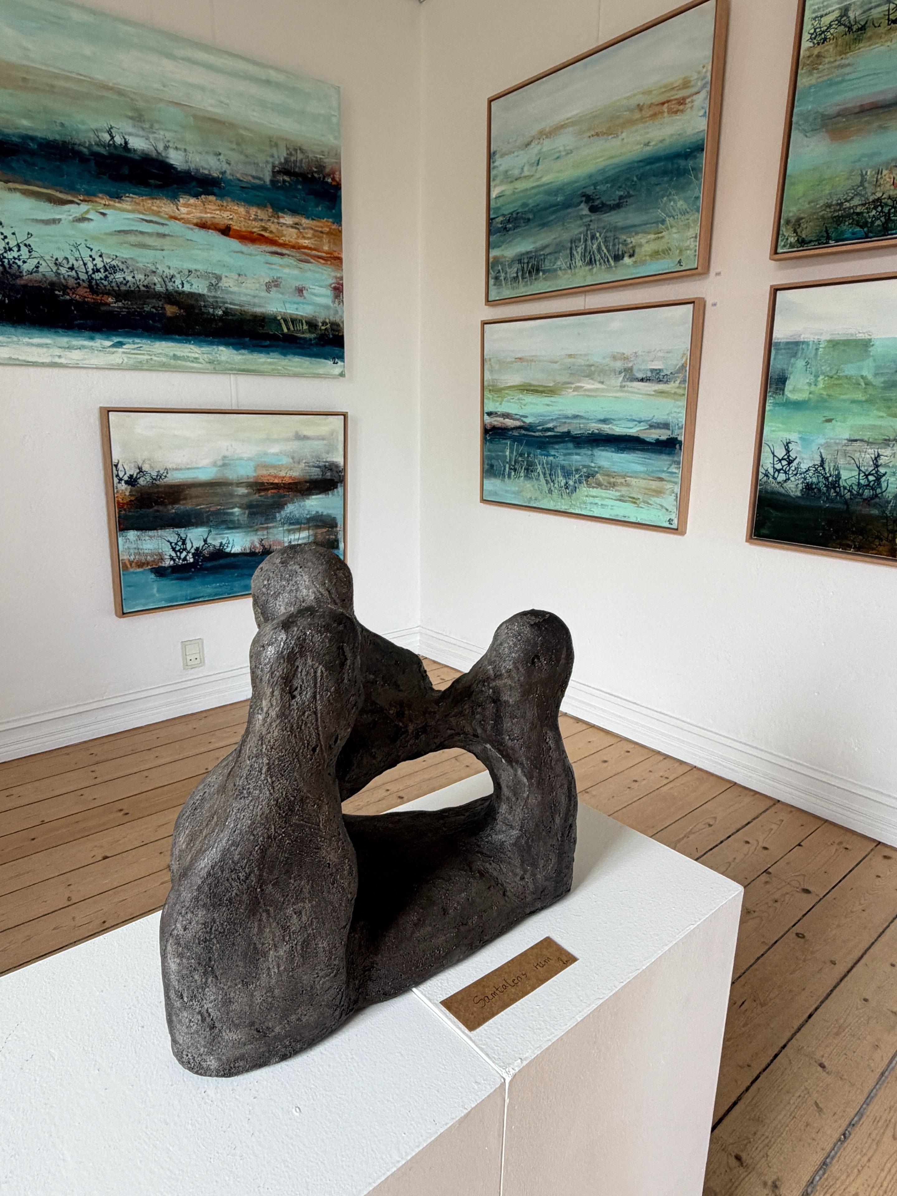 Skulptur af to figurer, der krammer, med malerier i blå og grønne nuancer på væggene i galleriet.