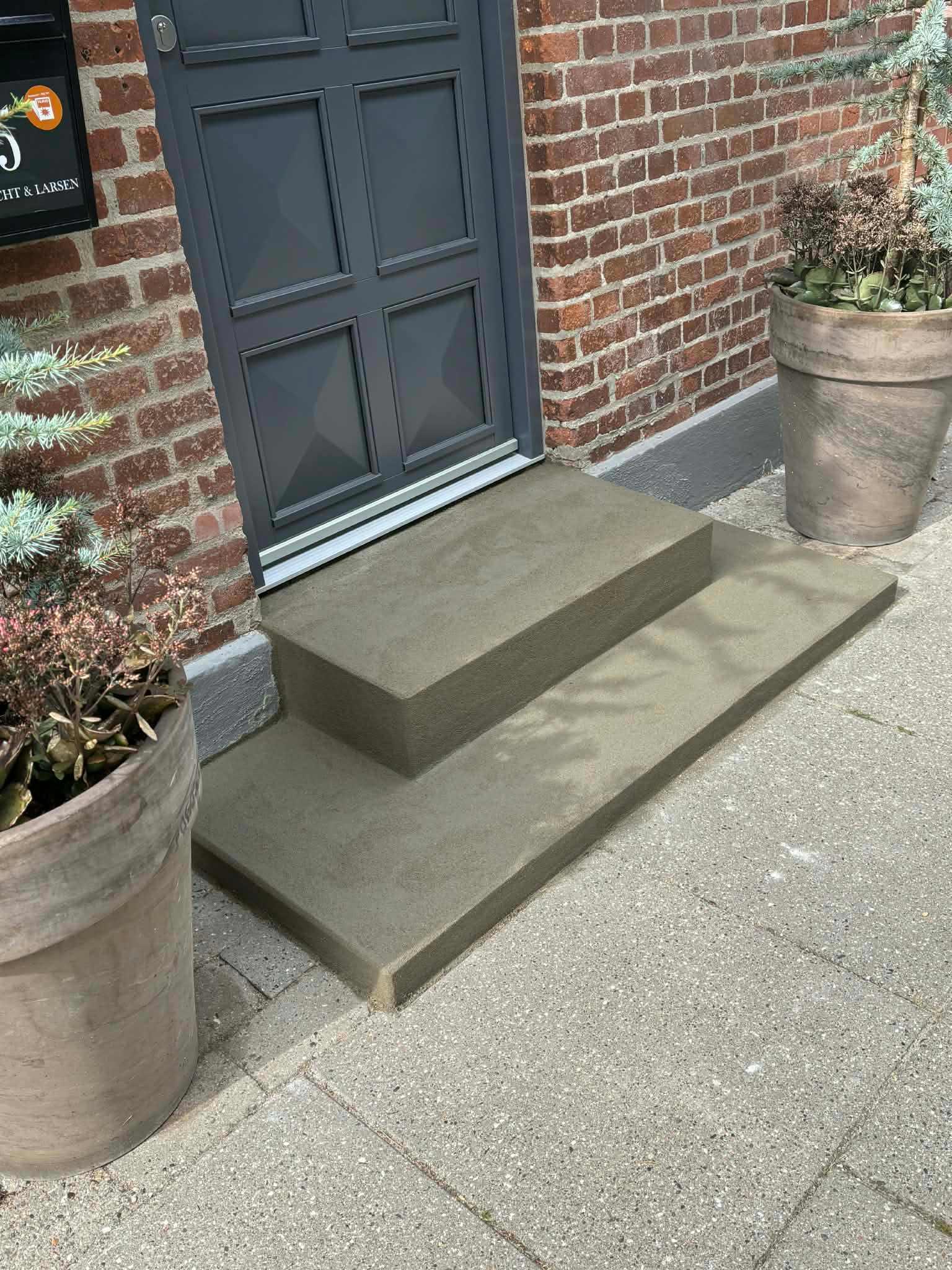 Trappe med to trin i grå beton foran en dør, omgivet af store plantekrukker.