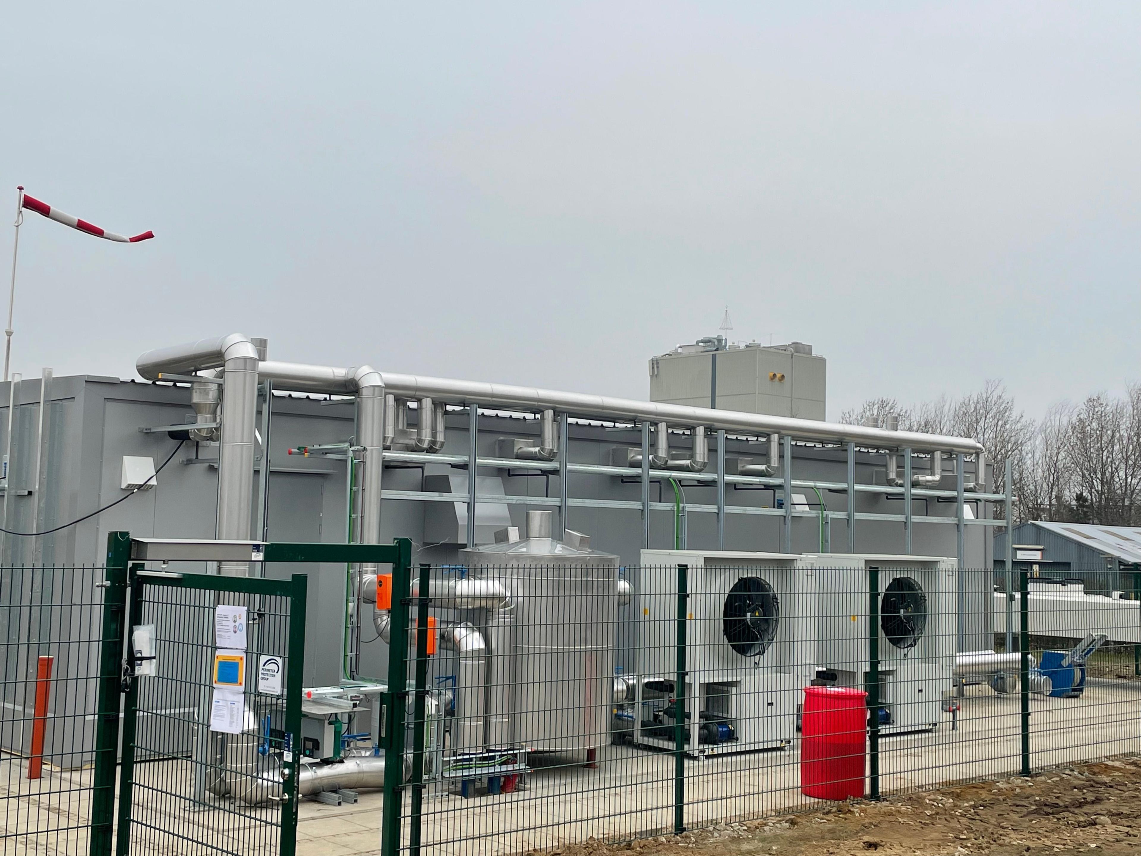 En stor industriel bygning med ventilationssystemer og maskiner, omgivet af et hegn og en grå himmel.