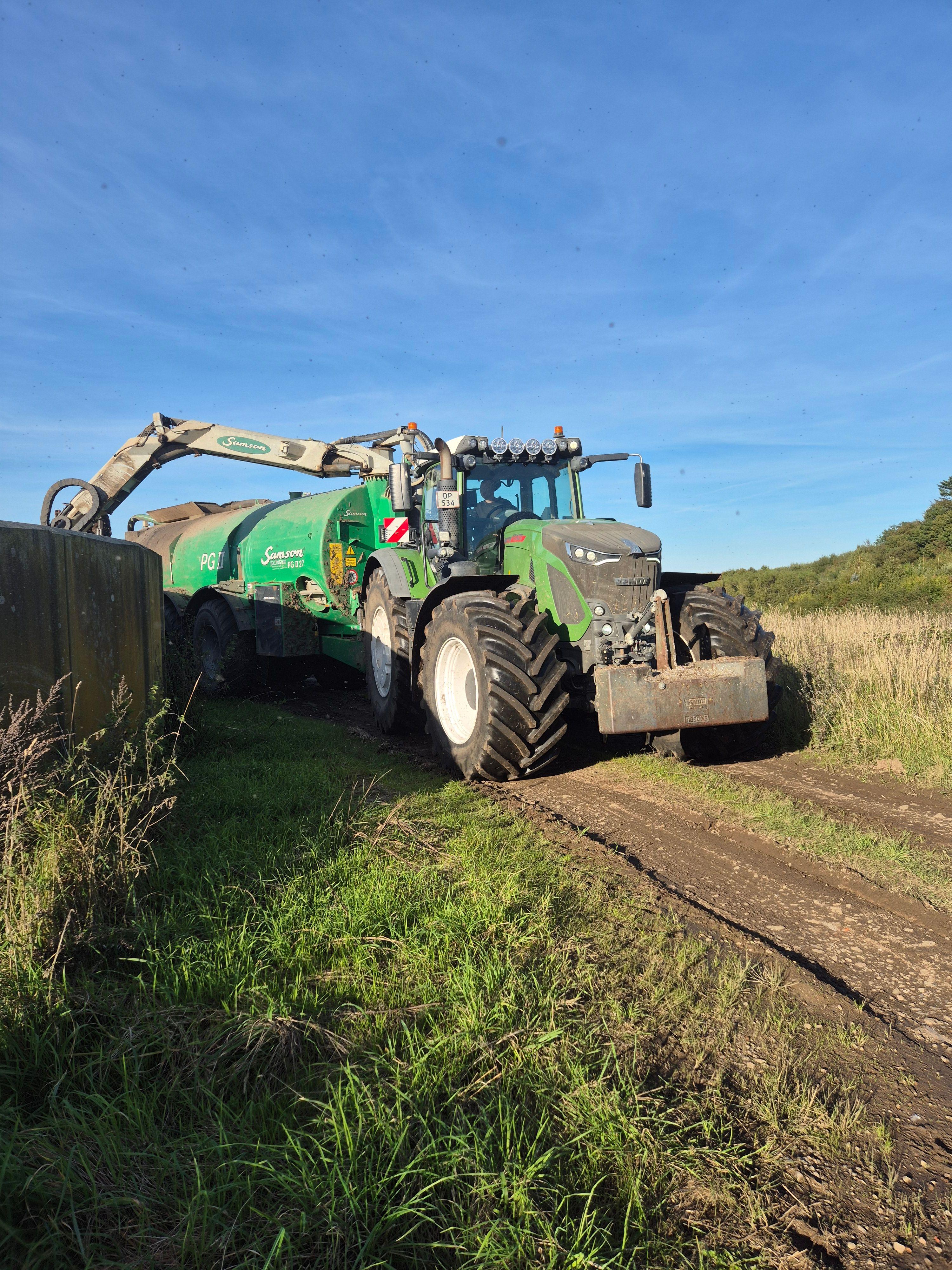 En stor traktor kører på en grusvej i et åbent landskab med blå himmel og grønne marker.