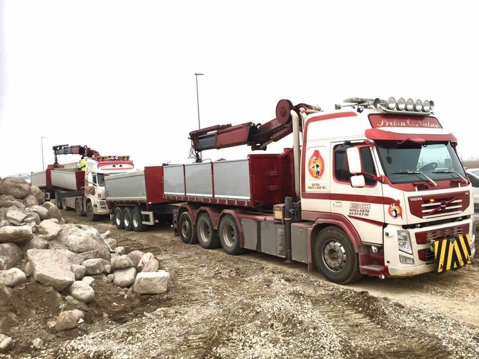 To store lastbil med kran og trailer, der transporterer materialer på en byggeplads. Sten og grus i baggrunden.