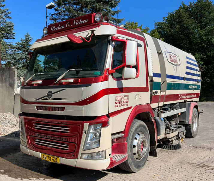 En rød og hvid fejeblad fra Volvo parkeret på en grusvej med træer i baggrunden.