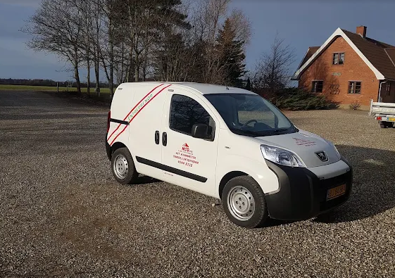 Hvid varevogn parkeret på grusvej med træer og hus i baggrunden. Varevognen har røde striber og logo.