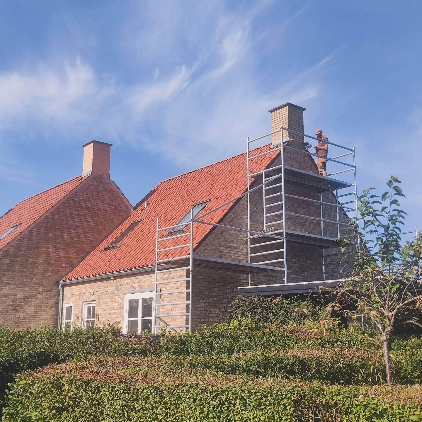 Hus med rødt tag og stillads, hvor en håndværker arbejder på skorstenen under en blå himmel.